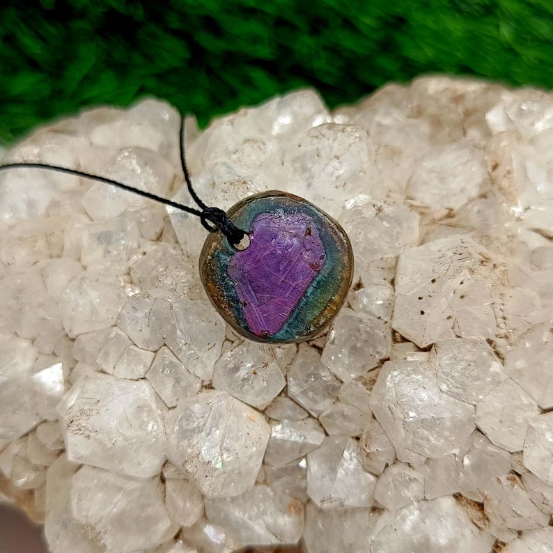 Pode incluir: Um pingente redondo com um centro roxo e iridescente, embutido em um metal de cor bronze. O pingente pende de um cord&atilde;o preto. O fundo &eacute; um aglomerado de cristais brancos, com folhagem verde vis&iacute;vel no canto superior esquerdo.