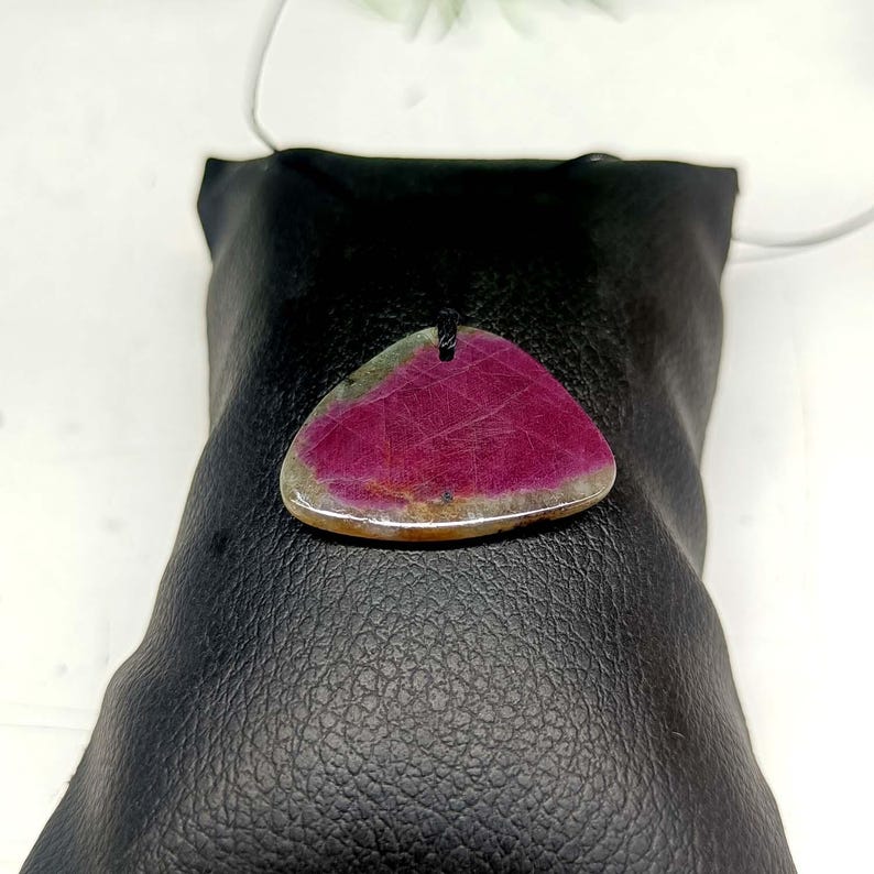 Pu&ograve; includere: Un ciondolo triangolare caratterizzato da un vivace centro rosa magenta con venature naturali e un bordo traslucido grigio-marrone. La pietra ha una superficie liscia e lucida ed &egrave; sospesa a un sottile cordino nero, mostrando un netto gradiente.