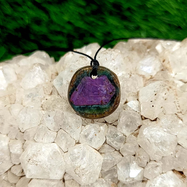 Pode incluir: Um pingente redondo com um design geom&eacute;trico roxo em uma moldura de cor bronze. O pingente &eacute; suspenso por um cord&atilde;o preto. O fundo apresenta cristais transparentes e folhagem verde.