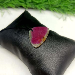 Pu&ograve; includere: Un ciondolo triangolare caratterizzato da una gemma color magenta vibrante e rosa intenso con inclusioni minerali naturali bianche e marrone chiaro, sospeso a un sottile cordino nero e appoggiato su un cuscino nero testurizzato.