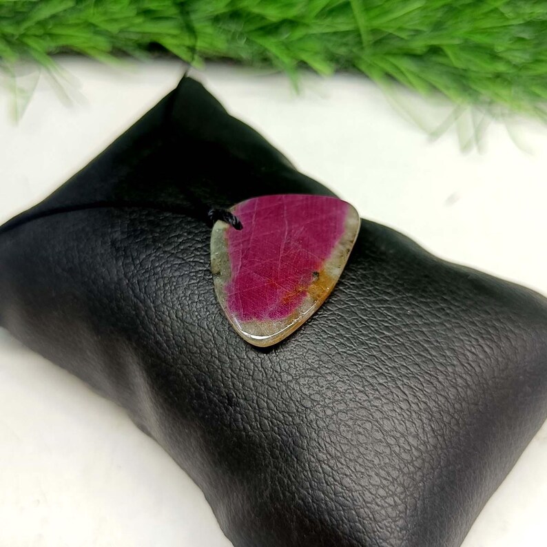 Pu&ograve; includere: Un ciondolo triangolare caratterizzato da una gemma color magenta vibrante e rosa intenso con inclusioni minerali naturali bianche e marrone chiaro, sospeso a un sottile cordino nero e appoggiato su un cuscino nero testurizzato.