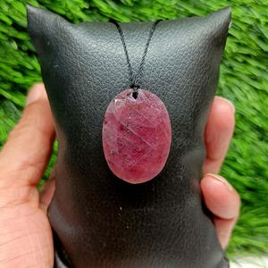 May include: An oval-shaped, faceted ruby pendant with a deep red hue and natural inclusions, suspended from a thin black cord. The gemstone features a polished surface with visible geometric facets that catch the light, showcasing a translucent quality.