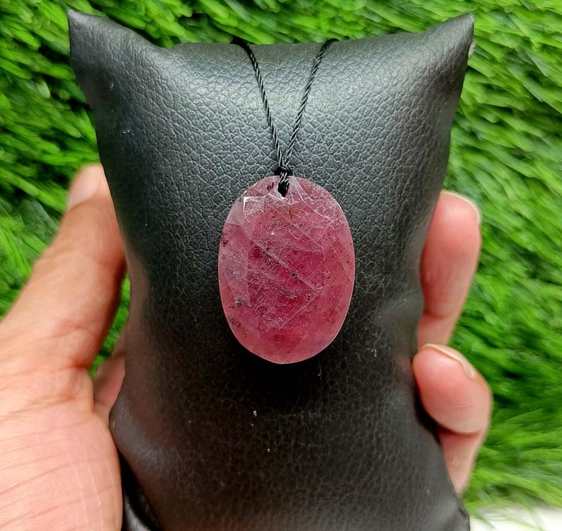 May include: An oval-shaped, faceted ruby pendant with a deep red hue and natural inclusions, suspended from a thin black cord. The gemstone features a polished surface with visible geometric facets that catch the light, showcasing a translucent quality.