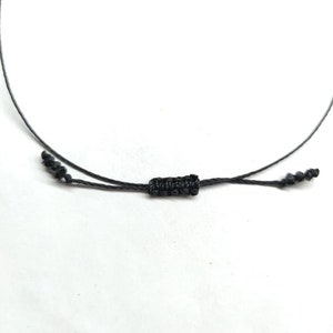 Pode incluir: Close-up de um cord&atilde;o de colar preto ajust&aacute;vel. O cord&atilde;o apresenta um n&oacute; tecido no centro e extremidades atadas para ajustar o comprimento. O fundo &eacute; branco.