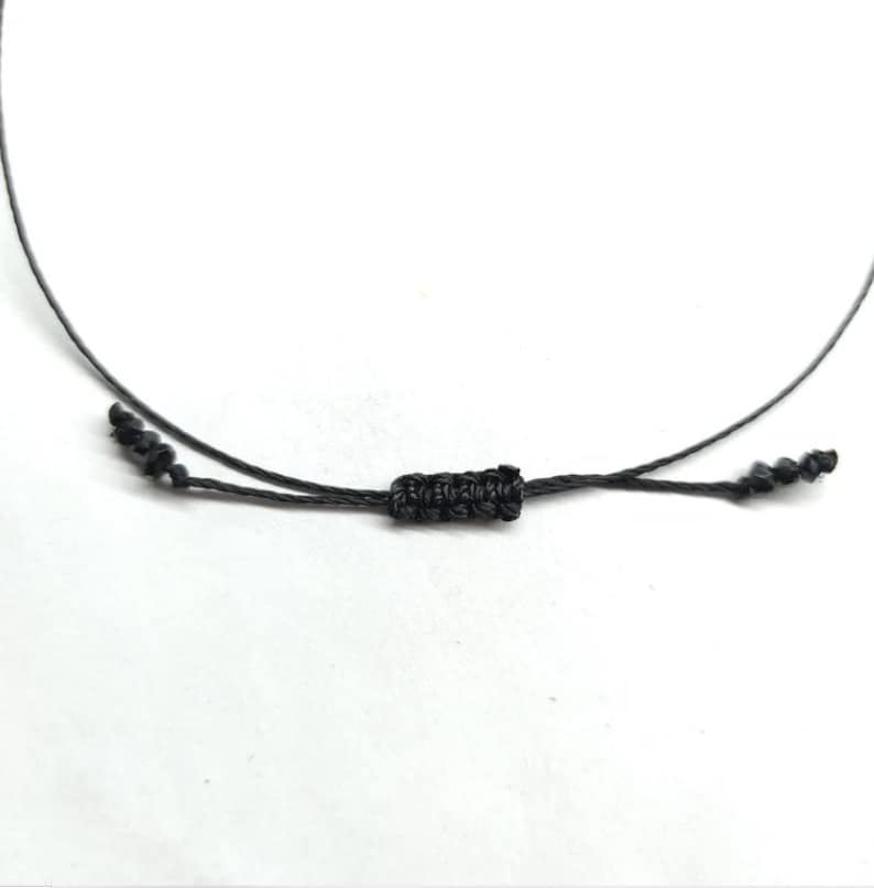 Pode incluir: Close-up de um cord&atilde;o de colar preto ajust&aacute;vel. O cord&atilde;o apresenta um n&oacute; tecido no centro e extremidades atadas para ajustar o comprimento. O fundo &eacute; branco.