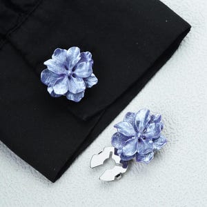 Könnte beinhalten: Ein Paar blaue, blütenförmige Manschettenknöpfe mit Metallic-Finish. Die Manschettenknöpfe sind vor einem schwarzen Hemdenärmel platziert und zeigen ihr aufwendiges Blumendesign und silberfarbene Metallverschlüsse. Das Bild hebt die eleganten Details der Manschettenknöpfe hervor.