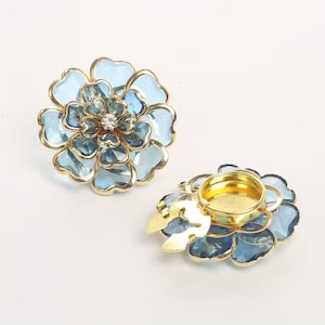 May include: Two flower-shaped candle holders with a gold-coloured rim and a clear gem in the centre. The petals are light blue. The candle holders are designed to resemble flowers.
