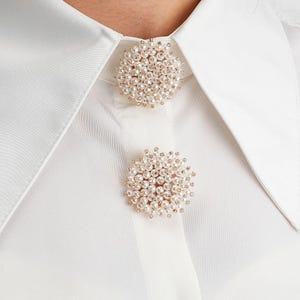 May include: Close-up of a white collared shirt with two pearl and gold-toned cluster brooches. The brooches are round and feature small, white pearls. A gold button is visible at the bottom of the shirt.