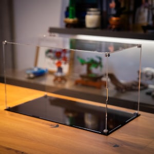 May include: A clear acrylic display case with a black base. The case is empty and sits on a wooden table.