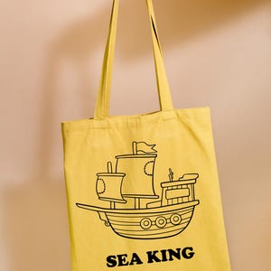 May include: A yellow tote bag with black outlines of a ship and the text "SEA KING".
