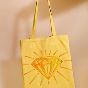 May include: A yellow canvas tote bag with a large, orange and yellow diamond graphic. The diamond is surrounded by sun rays.