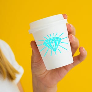 May include: A white to-go cup with a blue diamond graphic and a sunburst design. The cup is being held by a person's hand.
