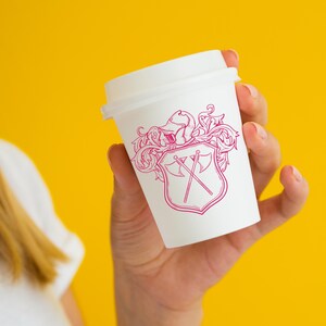 May include: A white to-go coffee cup with a pink and white design of a shield with crossed axes and floral accents.