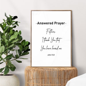 May include: A framed print with the text "Answered Prayer" in a bedroom setting. The print is propped on a woven side table next to a bed with white bedding. The text on the print reads "Father, I thank you that you have heard me, John 11:41."