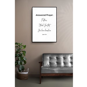 May include: A framed print with the text "~Answered Prayer~ Father, I Thank You that You have heard me, John 11:41." The black framed art hangs on a neutral wall above a dark gray leather couch. A potted plant in a woven basket sits to the left.