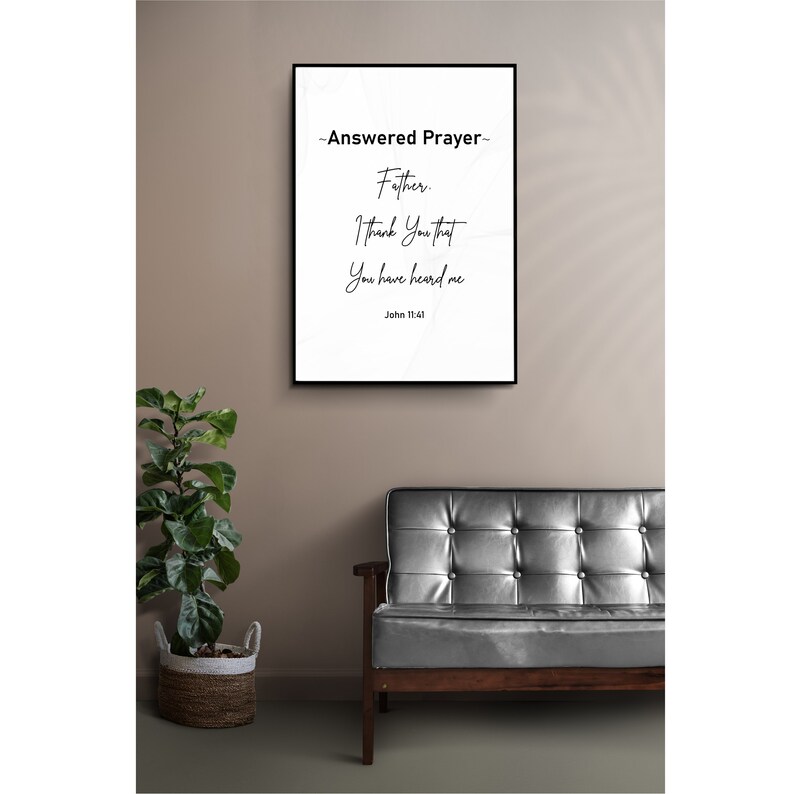 May include: A framed print with the text "~Answered Prayer~ Father, I Thank You that You have heard me, John 11:41." The black framed art hangs on a neutral wall above a dark gray leather couch. A potted plant in a woven basket sits to the left.