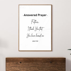 May include: A framed print with the words "Answered Prayer" and a Bible verse. The text is in black script on a white background, framed in a thin gold frame. The print is displayed above a wooden dresser.