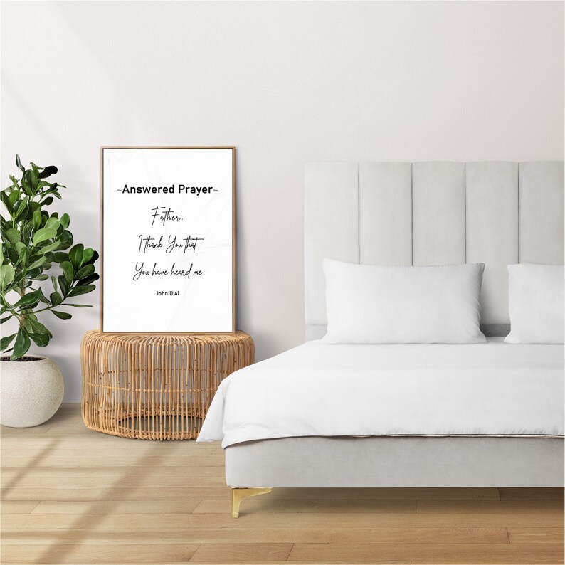 May include: A framed print with the text "Answered Prayer" in a bedroom setting. The print is propped on a woven side table next to a bed with white bedding. The text on the print reads "Father, I thank you that you have heard me, John 11:41."