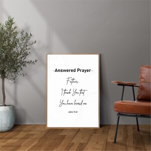 May include: A framed print with the words "Answered Prayer" and a Bible verse from John 11:41. The text is in black on a white background, framed in a thin, light brown frame. The print is displayed against a gray wall.
