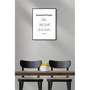 May include: A framed print with the text "-Answered Prayer- Father, I Thank You that You have heard me John 11:41." The print is in a black frame, hanging above a wooden table with two black chairs. A white bowl of yellow fruit sits on the table.