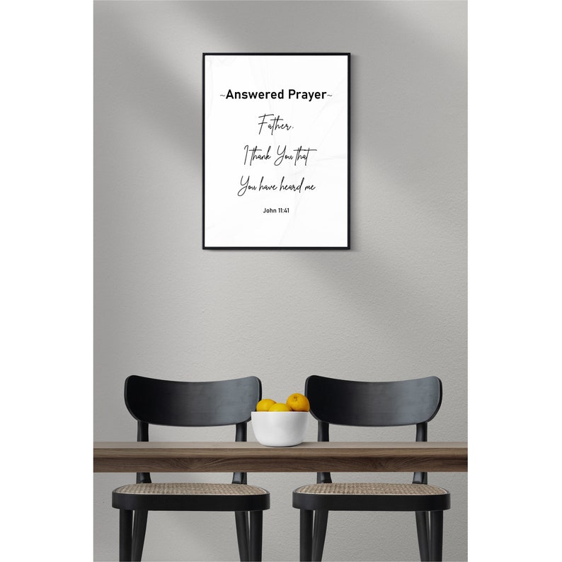 May include: A framed print with the text "-Answered Prayer- Father, I Thank You that You have heard me John 11:41." The print is in a black frame, hanging above a wooden table with two black chairs. A white bowl of yellow fruit sits on the table.