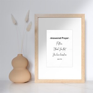 May include: A framed print with the text "-Answered Prayer- Father, I thank You that You have heard me John 11:41." The frame is light wood, and the print is white with black text. A beige vase with dried flowers is to the left.