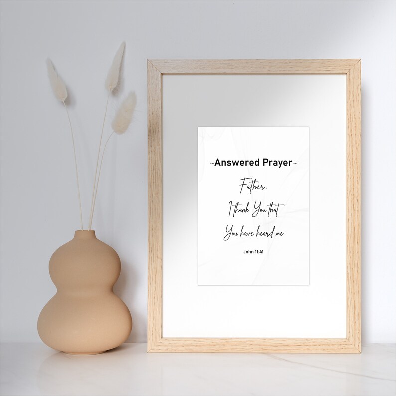 May include: A framed print with the text "-Answered Prayer- Father, I thank You that You have heard me John 11:41." The frame is light wood, and the print is white with black text. A beige vase with dried flowers is to the left.