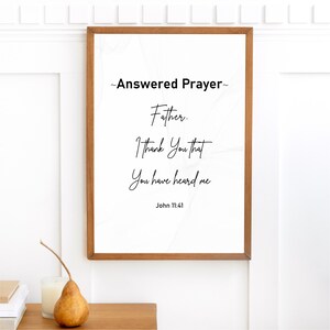 May include: A framed print with the text "Answered Prayer" and a quote from John 11:41. The print has a white background with a subtle pattern and is encased in a brown wooden frame. A pear and candle sit on a wooden surface.