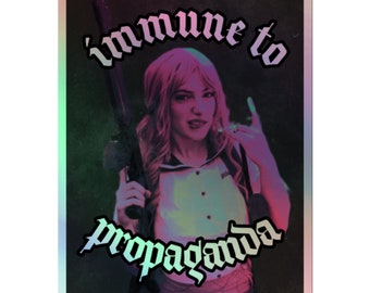 May include: A holographic sticker with a pink and blue gradient background. The sticker features a woman with pink hair holding a gun and making a hand gesture. The text "immune to propaganda" is written in black gothic font.