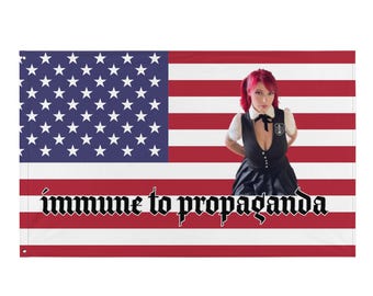 May include: A flag featuring the American flag design with the text "immune to propaganda" in a gothic font. A person with red hair and a black and white outfit is standing in front of the flag.