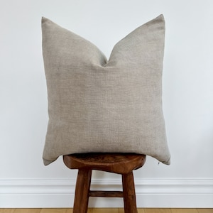 May include: A beige throw pillow with a subtle textured fabric. The pillow is sitting on a wooden stool.