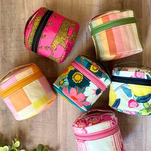 May include: Six small, cylindrical fabric pouches with zippered closures. The pouches feature various patterns including a pink leopard print, vertical pastel stripes, a yellow and pink checkerboard, a floral print, a fruit print, and a pink floral design.