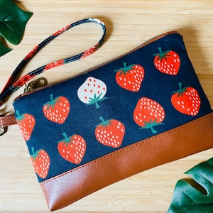 May include: A navy blue zippered pouch with a brown leather bottom and a wrist strap. The pouch is decorated with a pattern of red strawberries with green stems and leaves. The wrist strap has a red, white, and navy blue pattern.
