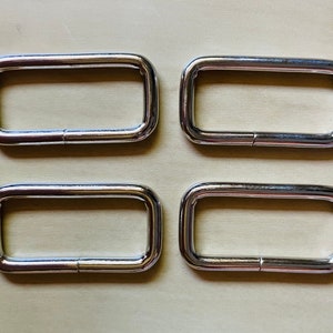 May include: Four silver metal D-rings, each with a rectangular shape and a smooth, polished finish.