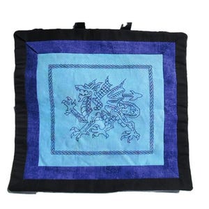 May include: Square quilted wall hanging featuring a blue dragon design. The dragon is embroidered in blue thread on a light blue background. The design is framed by a blue border, a darker blue border, and a black border. The wall hanging has a loop for hanging.
