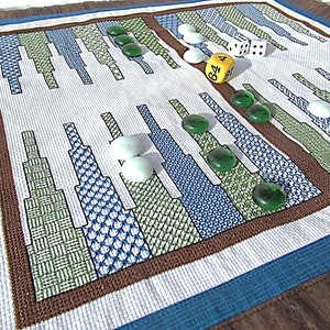 May include: A white fabric game board with a brown border and blue trim. The board has a pattern of green, blue, and white squares. There are white and green game pieces on the board, as well as two dice. One die shows the number 4 and the other die shows the number 6.