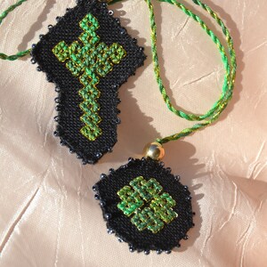Puede incluir: Dos colgantes de tela negra en forma de cruz con diseños bordados verdes, unidos a un cordón verde y dorado.