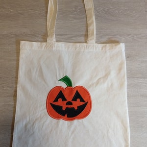 May include: A white canvas tote bag with a green stem and orange pumpkin face embroidered on the front.