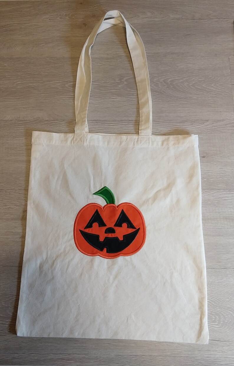 May include: A white canvas tote bag with a green stem and orange pumpkin face embroidered on the front.