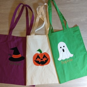 May include: Three Halloween-themed tote bags with different designs. The first bag is purple with a witch's hat and the name "Grace" embroidered on it. The second bag is white with a pumpkin and the third bag is green with a ghost and the name "Sophie" embroidered on it.