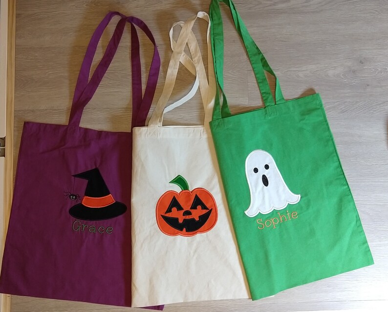 May include: Three Halloween-themed tote bags with different designs. The first bag is purple with a witch's hat and the name "Grace" embroidered on it. The second bag is white with a pumpkin and the third bag is green with a ghost and the name "Sophie" embroidered on it.