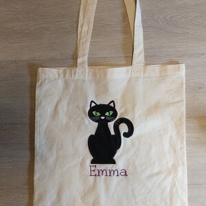 May include: White canvas tote bag with a black cat with green eyes and a pink nose. The cat is sitting and the name "Emma" is embroidered below it in purple thread.