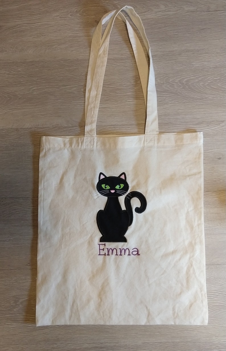 May include: White canvas tote bag with a black cat with green eyes and a pink nose. The cat is sitting and the name "Emma" is embroidered below it in purple thread.