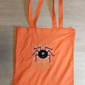 May include: Orange canvas tote bag with a black spider and web design and the name "Clara" embroidered in yellow thread.