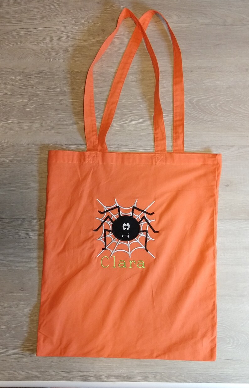 May include: Orange canvas tote bag with a black spider and web design and the name "Clara" embroidered in yellow thread.