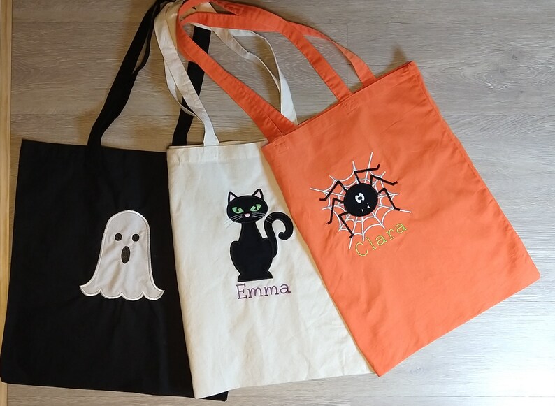 May include: Three Halloween-themed tote bags. One black bag with a white ghost, one white bag with a black cat, and one orange bag with a black spider.