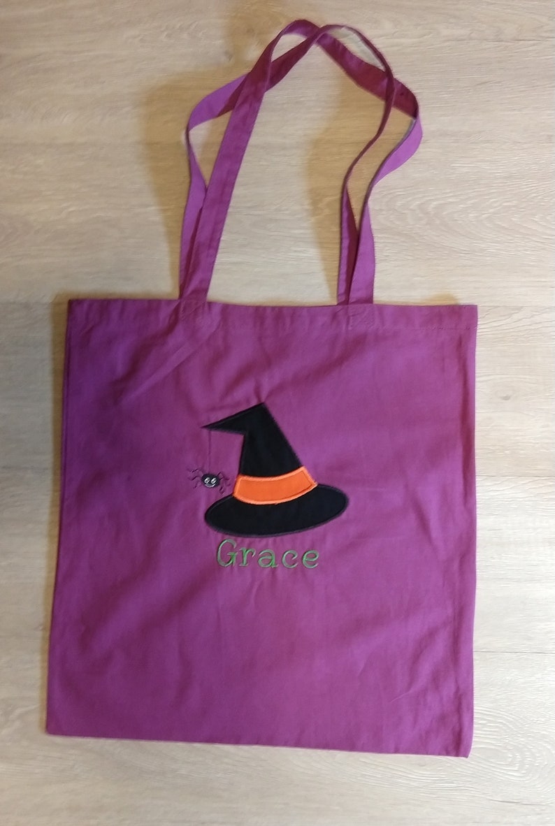 May include: Purple tote bag with a black witch hat and a spider embroidered on the front. The bag has the name "Grace" embroidered below the hat.