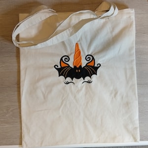 May include: A white canvas tote bag with an embroidered design of a black bat with orange and black wings and a curled orange horn. The bat has a black face with two white eyes.