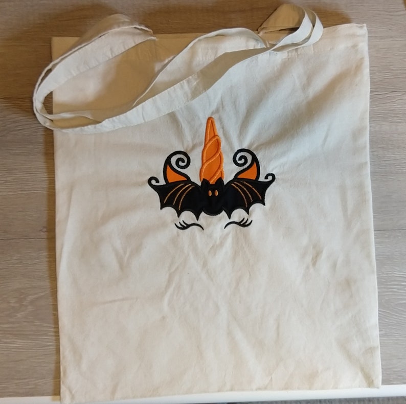 May include: A white canvas tote bag with an embroidered design of a black bat with orange and black wings and a curled orange horn. The bat has a black face with two white eyes.