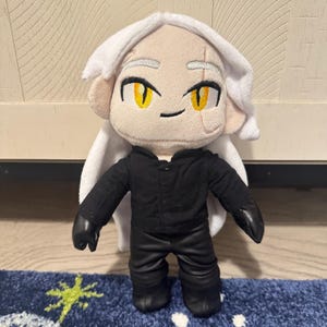May include: A plush doll with a light skin tone, white hair, and yellow eyes. The doll is wearing a black outfit with black gloves and boots. The doll has a stitched face and a white cape.